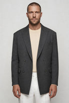 Man wearing a gray blazer over a beige shirt and white pants on a plain background